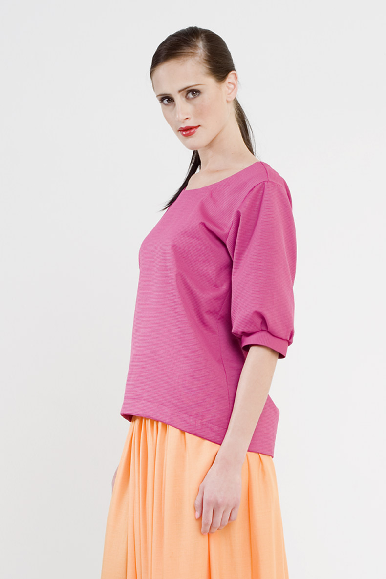 A woman with a shy smile is wearing her self-sewn pink shirt made from a sewing pattern.