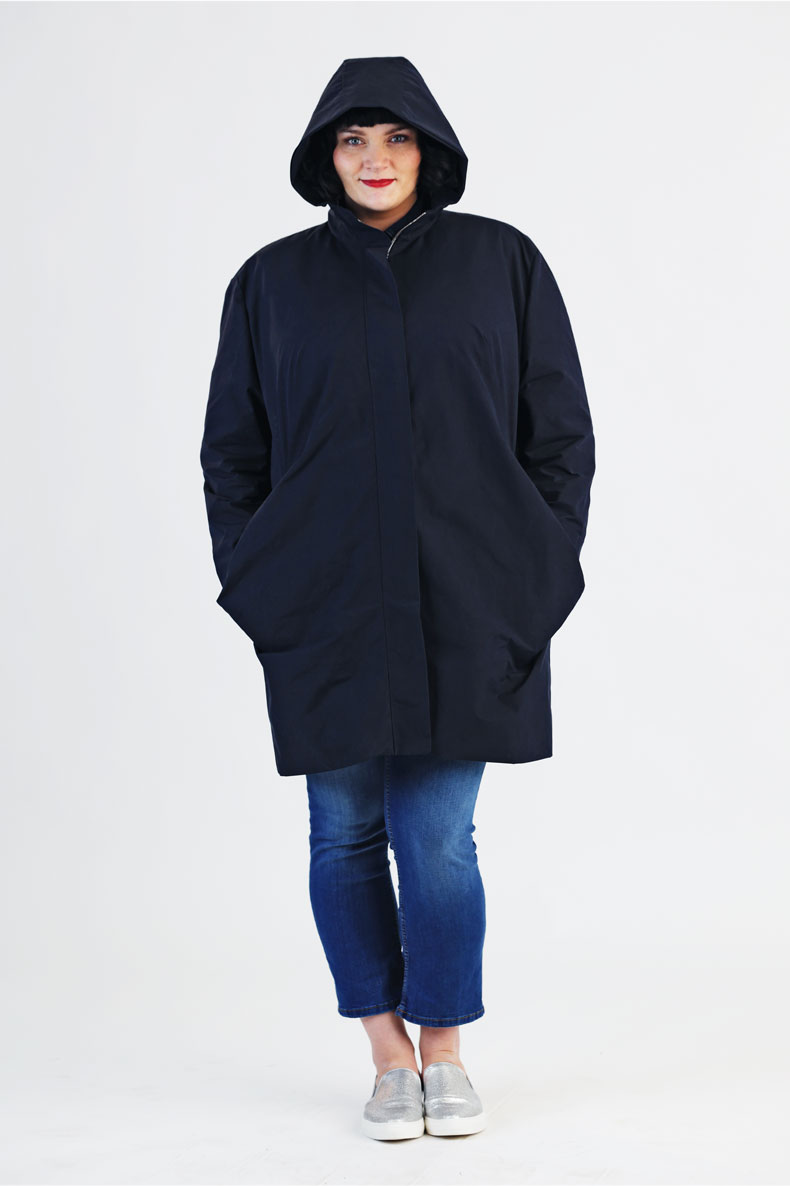 In front of a white wall, a woman is standing, wearing a blue plus-size parka.