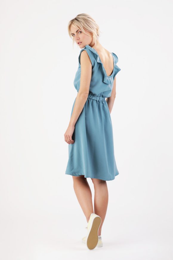 The back view of a woman standing in front of a white wall and wearing a blue dress.