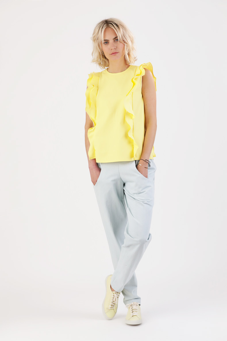 In front of a white wall, a woman is standing, wearing a yellow shirt and light gray pants.