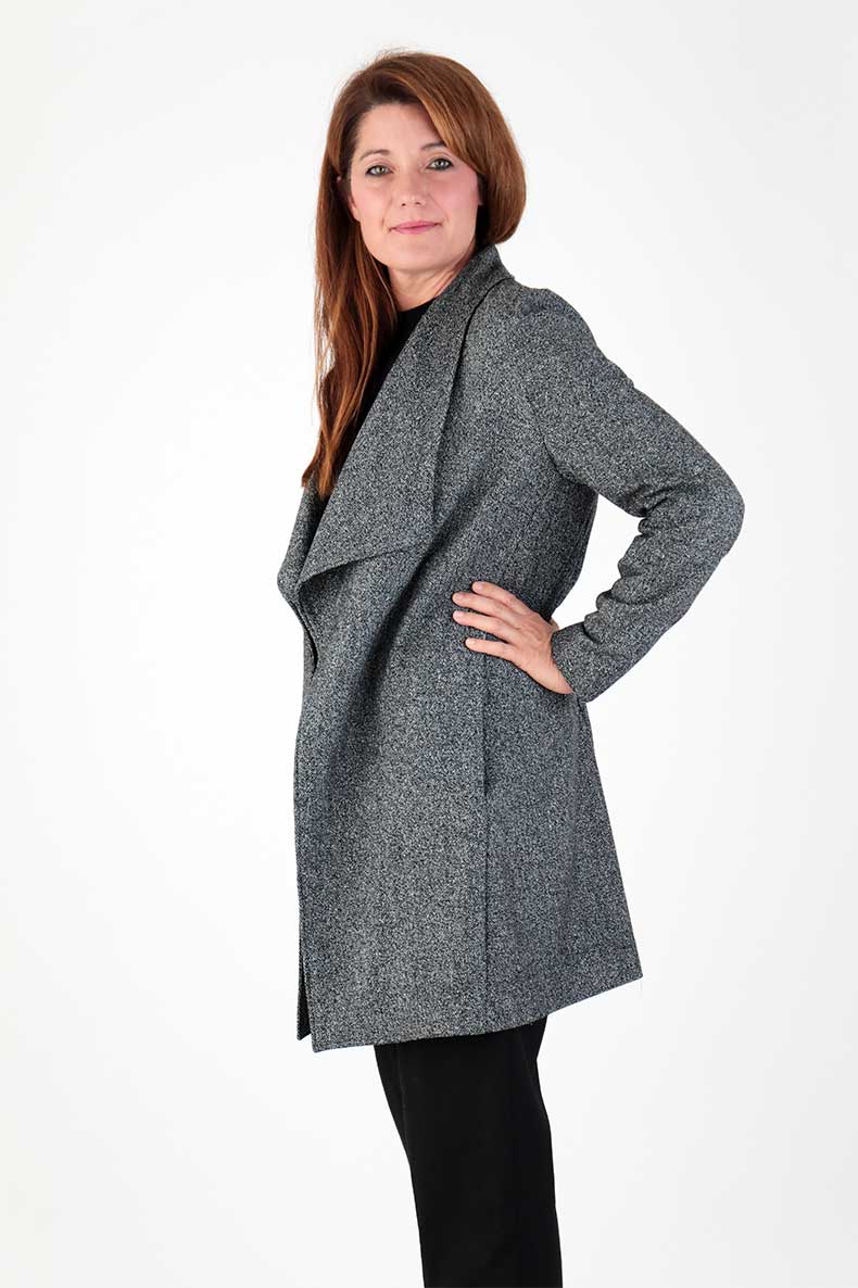 In front of a white background, a woman is standing with a gray coat and her hands on her hips.