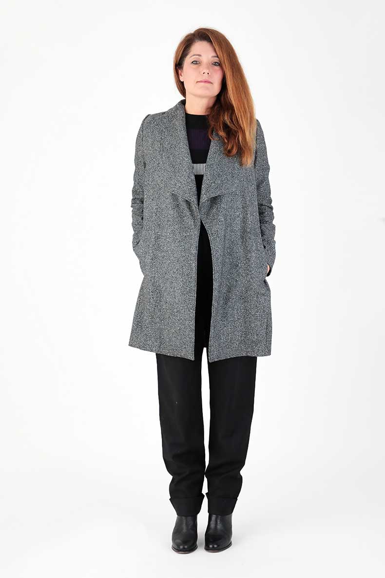 In front of a white wall, a woman is standing in her self-sewn coat.