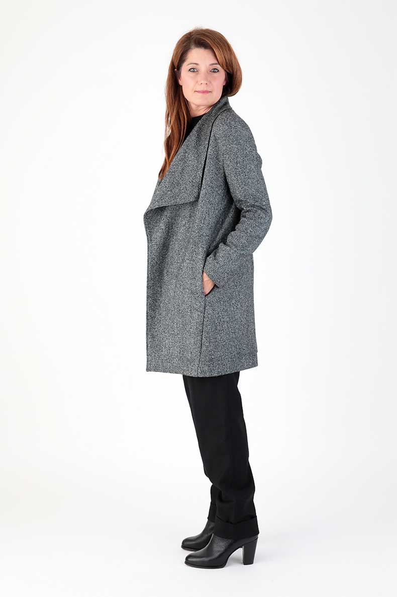 A woman is standing in front of a white background, hiding her hands in the pockets of her coat.