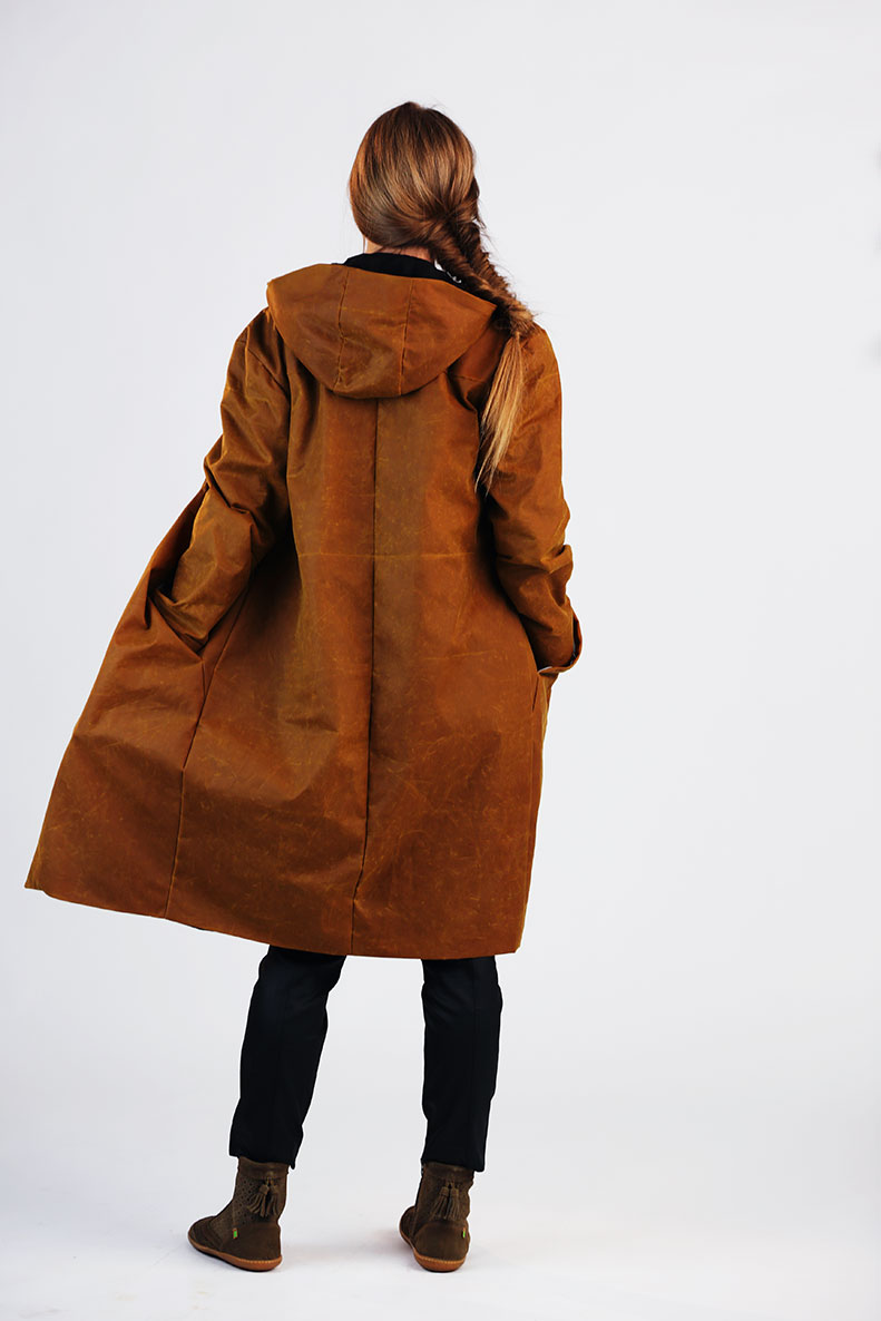 In front of a white background, a woman with a long braid is standing, and she is hiding her hands in the pockets of her brown anorak.