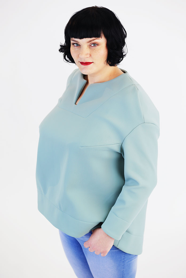 In front of a white background, a woman is standing in her self-sewn turquoise-colored shirt.