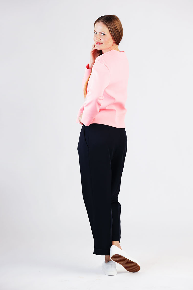 A woman with her back against the wall is wearing black pants and a pink shirt. She is smiling over her shoulder towards the camera.