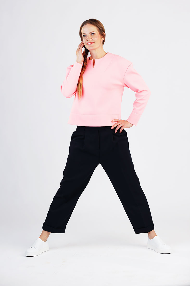 In front of a gray-white background, a smiling woman is standing, wearing black pants and a pink shirt.